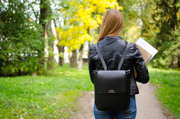 Вид сзади на молодую старшеклассницу или студентку колледжа, идущую в парк по дороге на занятия с рюкзаком и с учебниками в руках. Вернемся к школьной концепции. Вид сзади. Копирование пространства
. - Фото, изображение