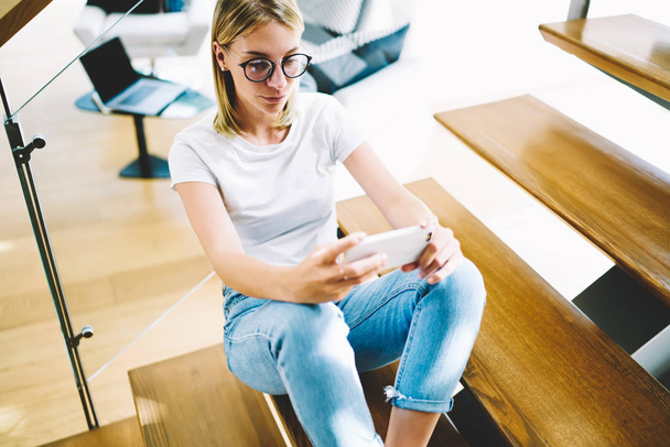 Young beautiful blonde woman in stylish eyeglasses reading news in social network using telephone with wireless connection. Gorgeous hipster girl making selfie on modern cellphone camera indoors - Photo, Image