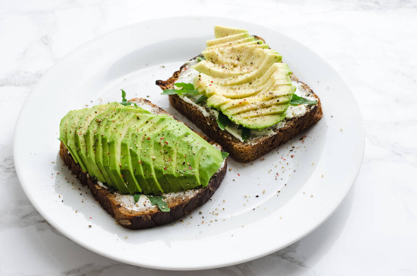 Avocado toasts on rye bread with sliced avocado, arugula, creme cheese with salt and pepper for healthy breakfast on light marble background. Vegetarian food. Plant-based diet. Clean eating concept. - Фото, зображення