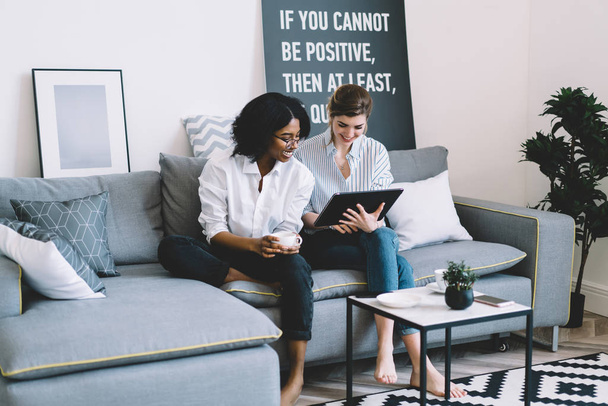Happy young diversity women choosing design clothes on web stores doing shopping online on modern touch pad device connected to home internet resting on comfortable couch in stylish apartment - Photo, Image