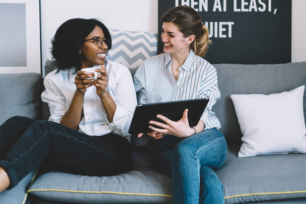 Happy african american young woman with coffee in hands laughing together with best caucasian friend during watching funny movie on modern touch pad relaxing on cozy sofa in stylish home interior - Foto, afbeelding
