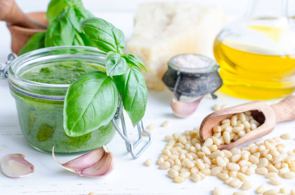 Pesto genovese - traditional Italian green basil sauce with raw ingredients on white wooden background. Basil leaves in mortar, Parmesan cheese, pine nuts, olive oil, garlic and salt. - Photo, Image