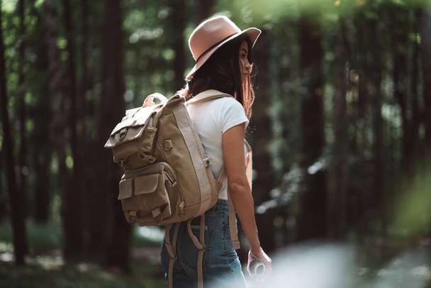 Back view on cute young woman with hat, backpack and location map in hand among trees in forest at sunset. - Φωτογραφία, εικόνα