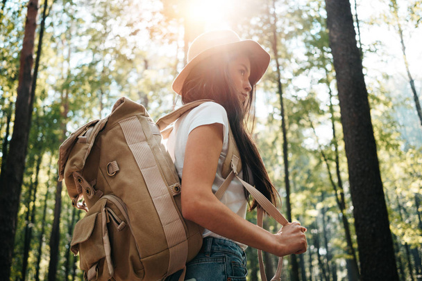 Young traveler woman wearing backpack and hat in hand among trees at sunset. Hipster girl with traveling backpack in forest. Lens flare effect - Foto, immagini