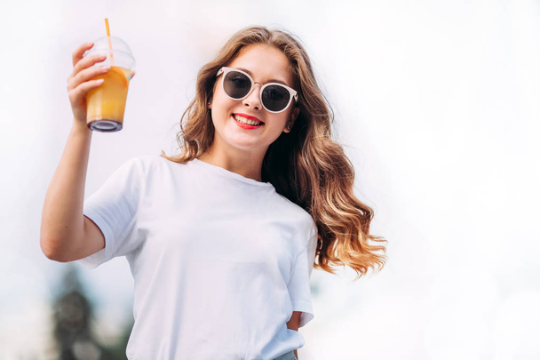 Close-up portrait of cheerful white woman in glasses on blur background. Photo of fashionable girl with beautiful brown hair smiling to camera and raises cocktail. - Фото, зображення