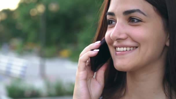 Happy girl is smiling and chatting on phone with her Mr. Right in summer                                            Portrait of a cheerful and stylish young woman with sparkling eyes talking on smartphone with her boyfriend on a riverbank in summer  - Séquence, vidéo