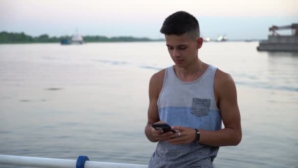 Young man stands and touches his phone at a river quay at sunset in summer                      Portrait of a young man with a crew hairstyle in a singlet standing and touching the screen of his smartphone on a embankment with handrails at sunset  - Video
