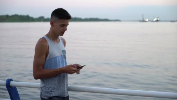 Sportive man stands and browses the net at a river quay at sunset in summer                      Profile of an athletic man in a white singlet standing and touching the screen of his smartphone on a embankment with handrails at a beautiful sunset  - Filmati, video