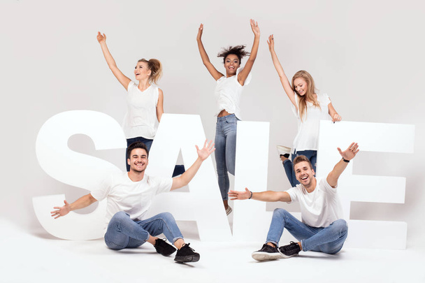 Group of young multi-ethnic beautiful people wearing jeans posing together with big sale letters against white background. Cheerfull smiling friends. - Photo, Image