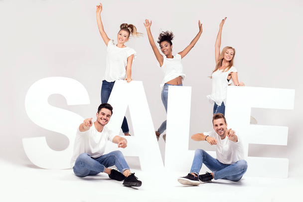 Group of young multi-ethnic beautiful people wearing jeans posing together with big sale letters against white background. Cheerfull smiling friends. - Photo, Image