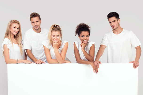 Ryhmä nuoria monietnisiä kauniita ihmisiä valkoisissa paidoissa, poseeraamassa tyhjän valkoisen laudan kanssa. Iloisia hymyileviä ystäviä
. - Valokuva, kuva