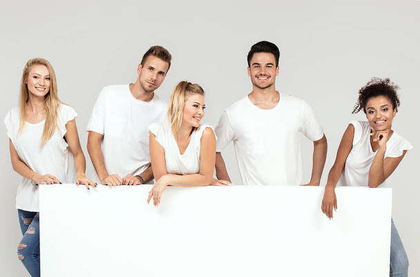 Group of young multi-ethnic beautiful people wearing white shirts, posing together with empty white board. Cheerfull smiling friends. - Foto, afbeelding