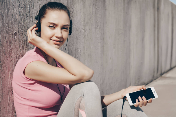 nice slender girl in a pink shirt resting after a long workout, listening to music on headphones - Photo, image