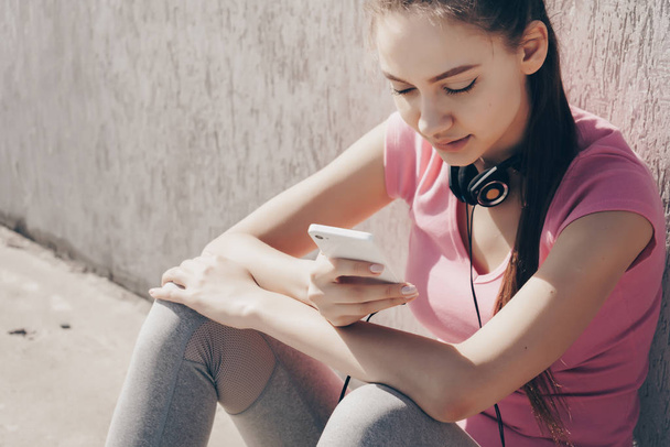 beautiful slender girl in a pink t-shirt sits on the ground, looks into her smartphone, neck hangs on her neck - Zdjęcie, obraz