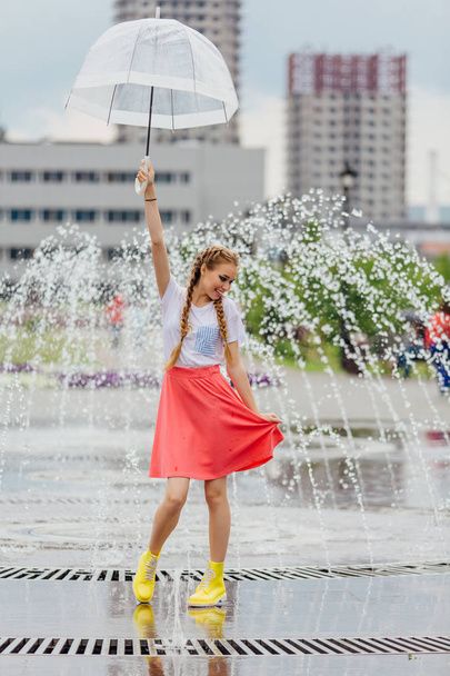 Young pretty girl with two braids in yellow boots and with transparent umbrella stands near fountain. Rainy day in city. - Photo, Image
