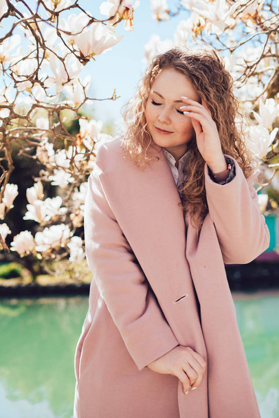 beautiful curly woman in a pink coat enjoying the scent of magnolia in the park - Fotó, kép