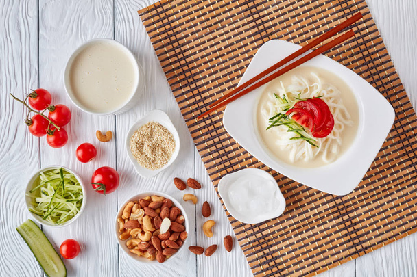 soy milk korean soup kongguksu, kong-guksu with rice noodle, nuts, and sesame topped with chopped cucumber, tomato in a white bowl with ingredients on a table, view from above, flat lay - Фото, зображення