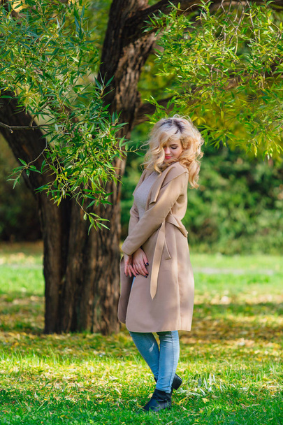 Beautiful elegant blonde woman dressed in a coat standing under the tree in autumn park. Sunny october day. - Foto, immagini
