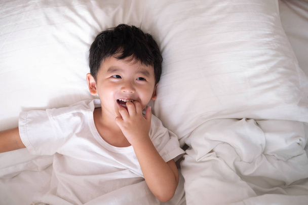 3 años de edad pequeño lindo chico asiático en camisa blanca en casa en la cama, niño acostado jugando y sonriendo en la cama blanca con almohada y manta, vista superior con espacio de copia para añadir texto
. - Foto, Imagen