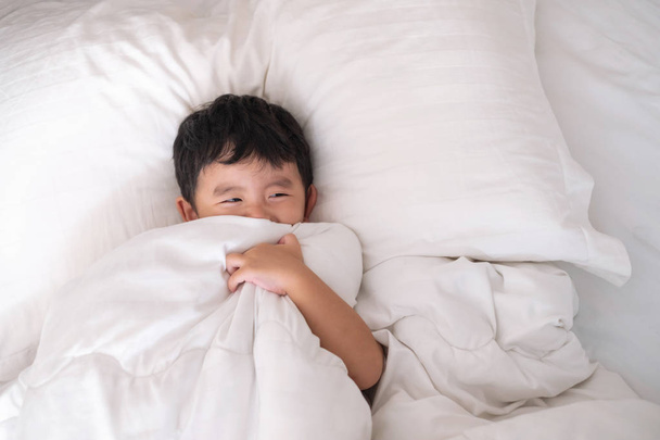 3 years old little cute Asian boy in white shirt at home on the bed, kid lying playing and smiling on white bed with pillow and blanket, top view with copy space for add text. - Photo, Image