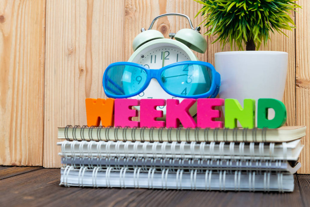 WEEKEND letters text and notebook paper, alarm clock and little decoration tree in white vase on wooden background, hello weekend vacation concept idea. - 写真・画像