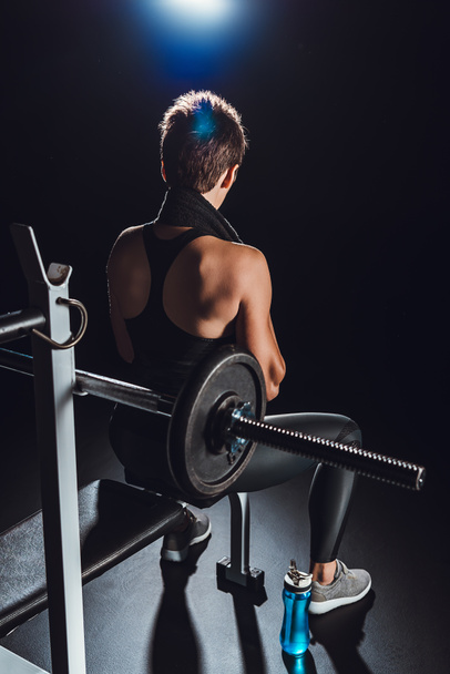 rear view of sportswoman with towel over neck resting near barbell at gym with bottle of water on floor, black background - Фото, изображение