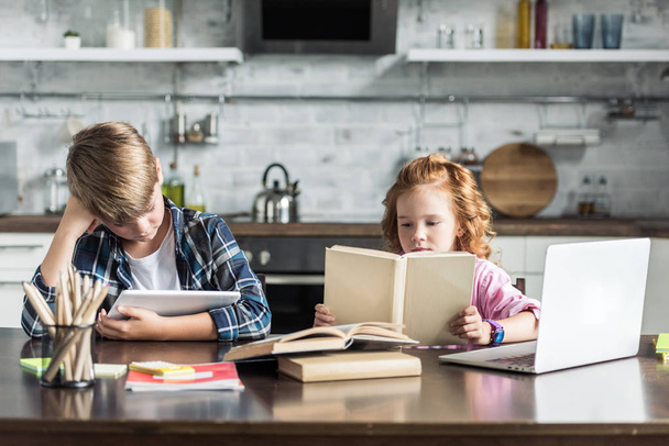 little brother and sister with laptop and tablet doing homework at kitchen - 写真・画像