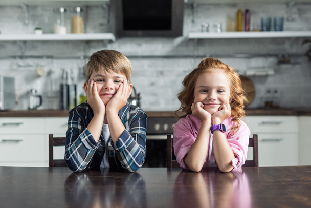 smiling little brother and sister sitting at kitchen together and looking at camera - 写真・画像