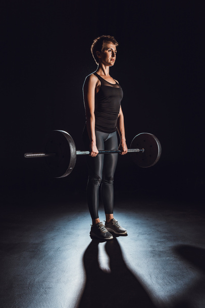 serious sportswoman exercising with barbell at gym, black background - Photo, Image