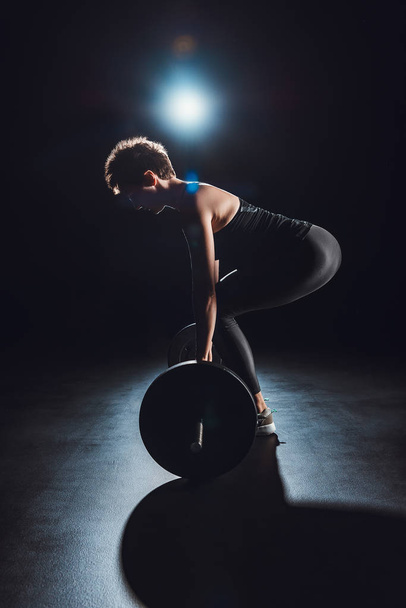 вид сбоку женщины, тренирующейся со штангой в тренажерном зале, черный фон
  - Фото, изображение