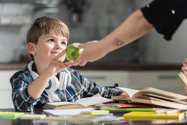 Schnappschuss von Mutter, die ihrem Sohn grünen Apfel gibt, während er Hausaufgaben macht - Foto, Bild