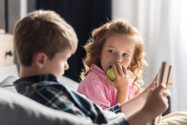 cute little schoolboy reading book on couch while his sister eating apple - Foto, Bild