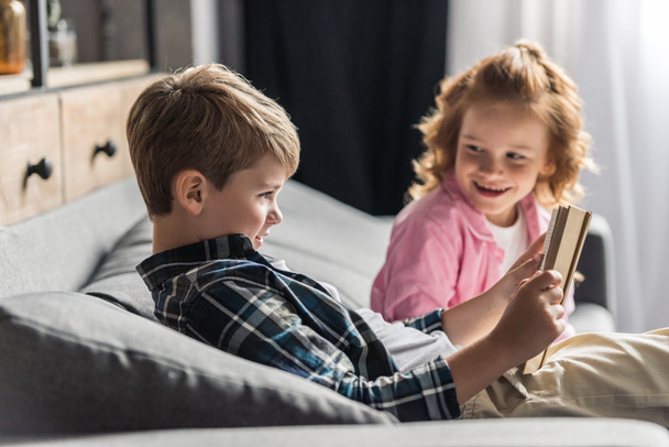 cute little schoolboy reading book on couch while his sister talking to him - Foto, Bild