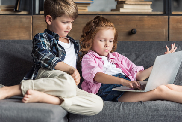 irmãozinho e irmã usando laptop juntos enquanto relaxa no sofá
 - Foto, Imagem