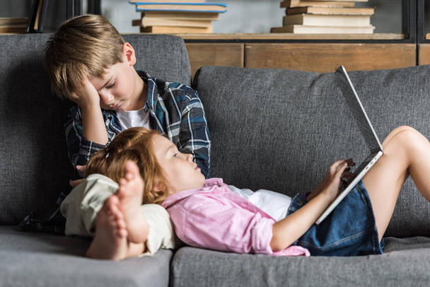 little girl lying with head on legs of brother and using laptop while they relaxing on couch - Foto, afbeelding