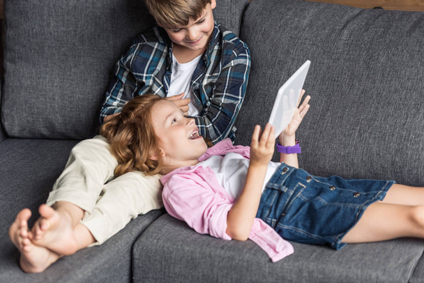 little brother and sister using tablet together while relaxing on couch - Foto, Bild