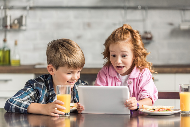 sorprendido hermano pequeño y hermana usando tableta durante el desayuno
 - Foto, Imagen