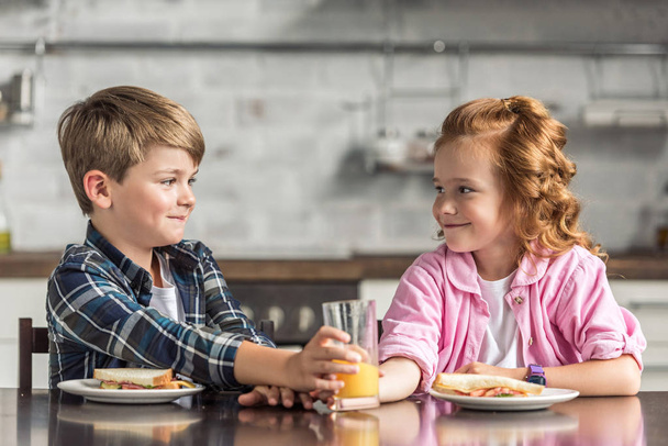 little brother and sister reaching for glass of orange juice during breakfast and looking at each other - Foto, afbeelding