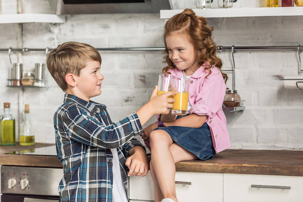 little brother and sister clinking glasses of orange juice at kitchen - Foto, Imagem