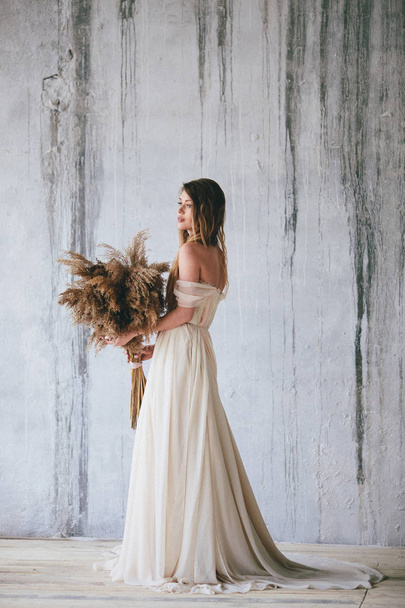 girl in a beige flying dress with the sheaves of hay in her hands on the old wall background. Wedding conception - Photo, Image