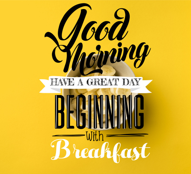 Bovenaanzicht van kom met gesneden bananen geïsoleerd op geel met "Goedemorgen, hebben een geweldige dag, beginnen met ontbijt" inspiratie - Foto, afbeelding
