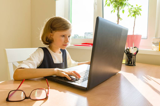 Little schoolgirl uses computer sitting at a desk at home. School, education, knowledge and children - Foto, Imagem