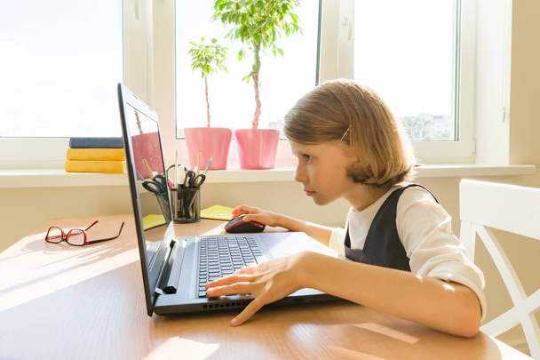 Little schoolgirl uses computer sitting at a desk at home. School, education, knowledge and children - Foto, immagini