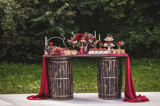 Table en bois de réception de mariage debout sur deux barils décorés de fleurs rouges et sucrées avec des boissons et de la soie, style rustique
  - Photo, image