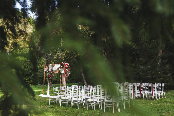 Cerimônia de casamento em estilo rústico decorado com flores vermelhas e têxteis com cadeiras brancas na floresta
 - Foto, Imagem