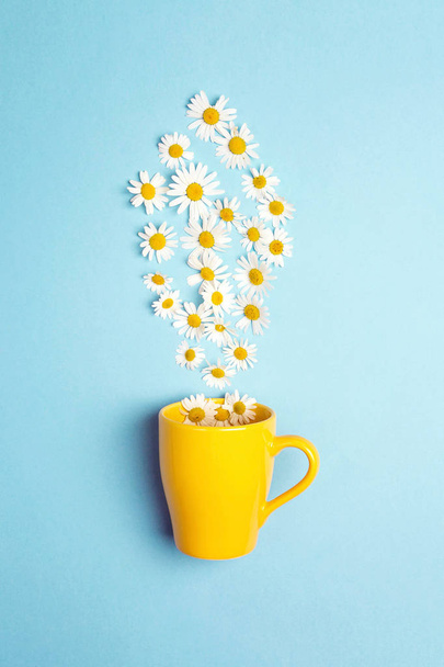 Yellow mug and chamomile flowers on a blue background. Chamomiles come out of the mug like steam. Chamomile tea concept. Flat lay, top view. - Фото, изображение