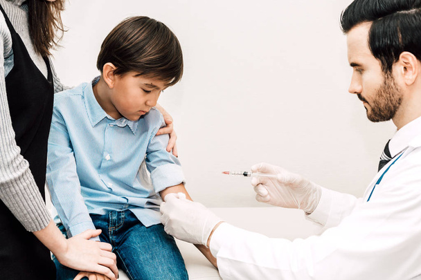 Lääkäri suojakäsineissä antaa injektio pienen pojan lapsen ja äidin kanssa lääkäreiden pöydällä sairaalassa.Terveydenhuolto ja lääketiede
 - Valokuva, kuva