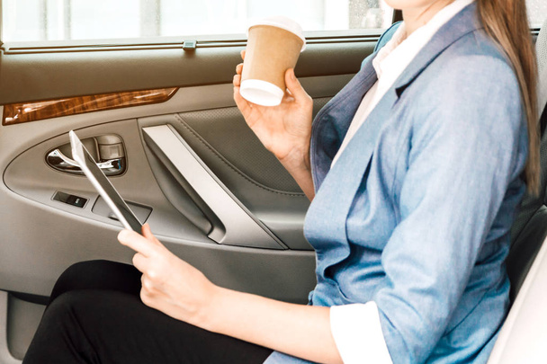 Liikenainen työskentelee digitaalisen tabletin tietokoneen kanssa ja pitää kahvia ylellisen auton takapenkillä
 - Valokuva, kuva