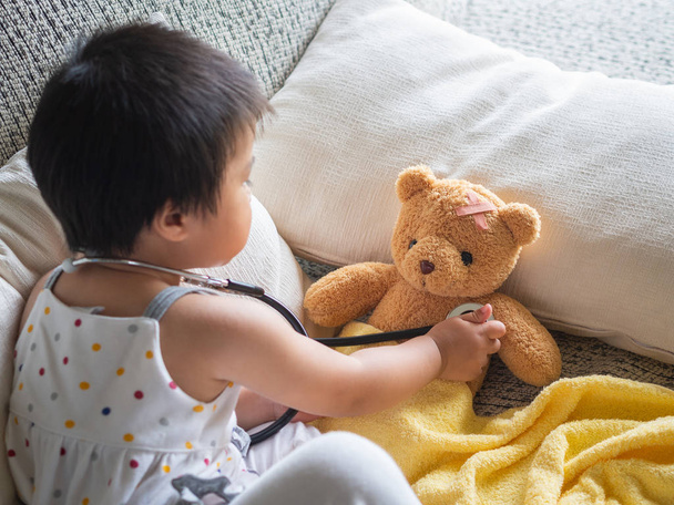Menina bonito pequeno feliz está jogando médico com estetoscópio e urso boneca. Conceito de criança e cuidados de saúde
. - Foto, Imagem
