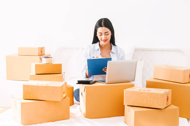 Young woman freelancer working and checklist and writing order with cardboard box on bed at home - SME business online and delivery concept - Fotó, kép